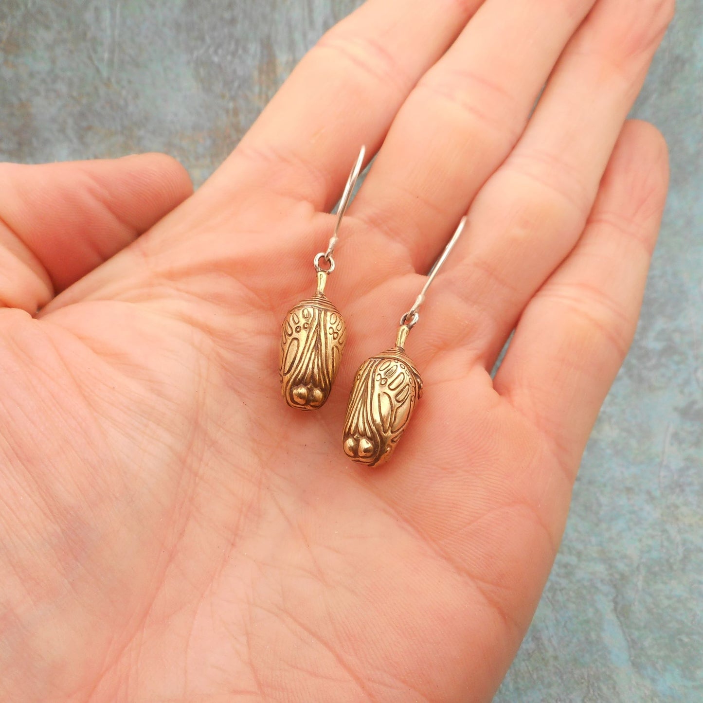 A photo of a pair of brass chrysalis earrings, hanging on the palm of a hand, on a blue background.