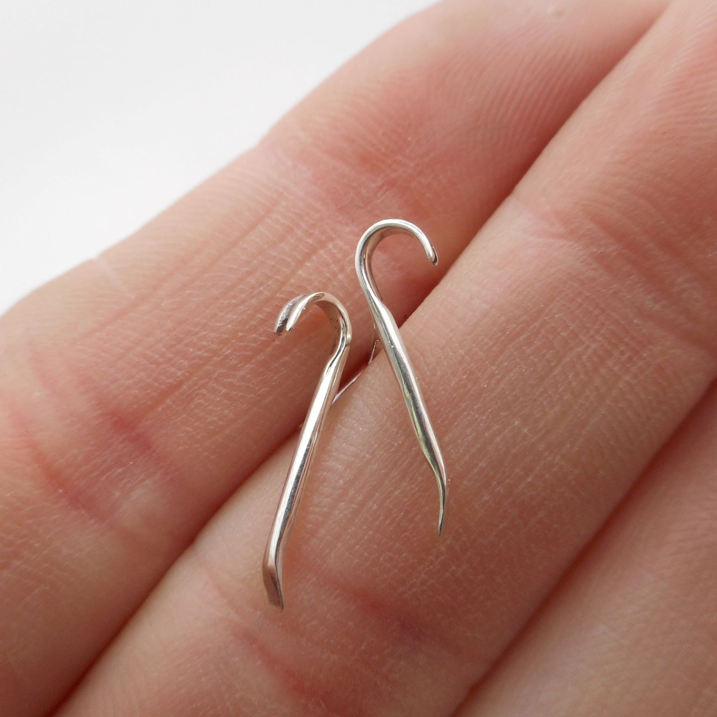 A photo of two miniature silver crowbar earrings, resting on the fingers of a hand.