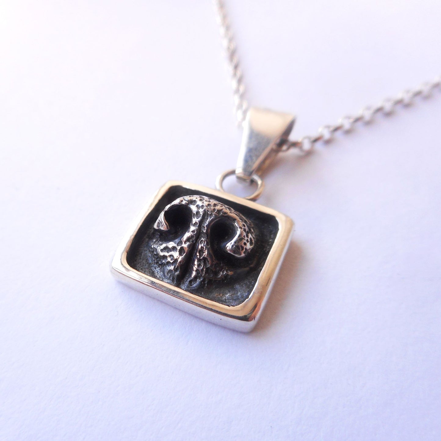 A photo of a silver pendant, shaped like a dog's nose in a square frame, on a white background.