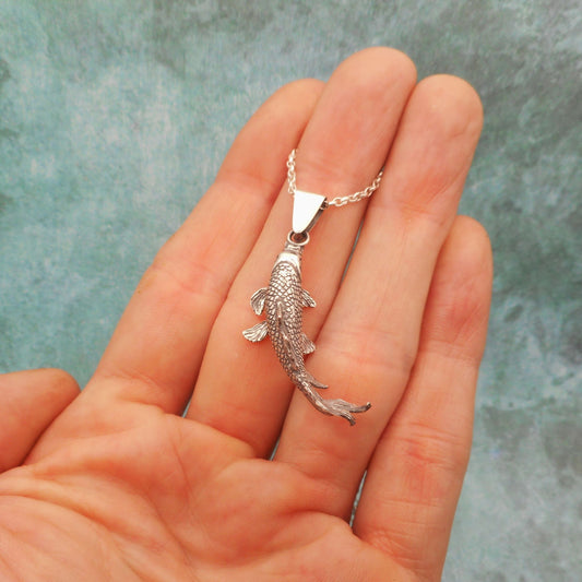 A photo of a silver koi fish pendant, resting on a hand, on a blue background.