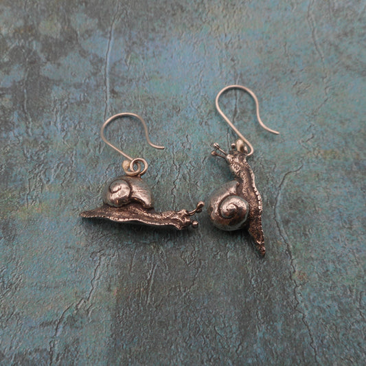 A photo of a two miniature silver snail earrings, on a mottled blue background.
