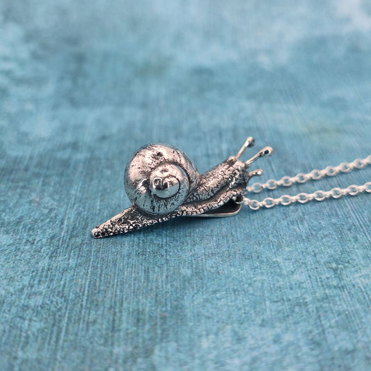 A photo of a silver pendant in the shape of a snail, on a blue background.