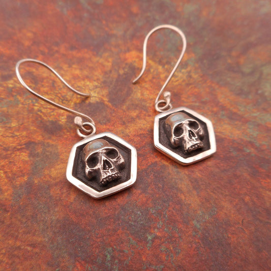 A photo of a pair of silver earrings, shaped like skulls in hexagonal frames, resting on a mottled red background.