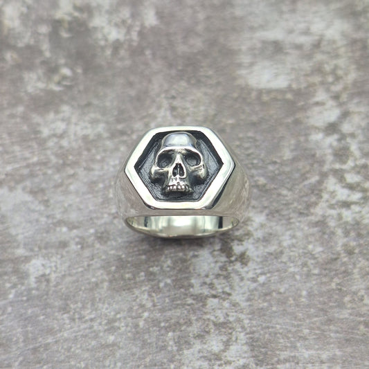 A photo of a silver signet ring with a small skull on the top surface, resting on a grey background.