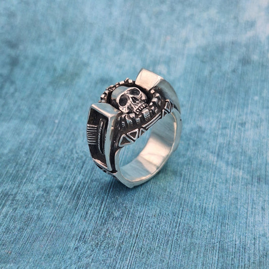 A photo of a silver ring, featuring a skull and some industrial shapes, resting on a mottled blue background.