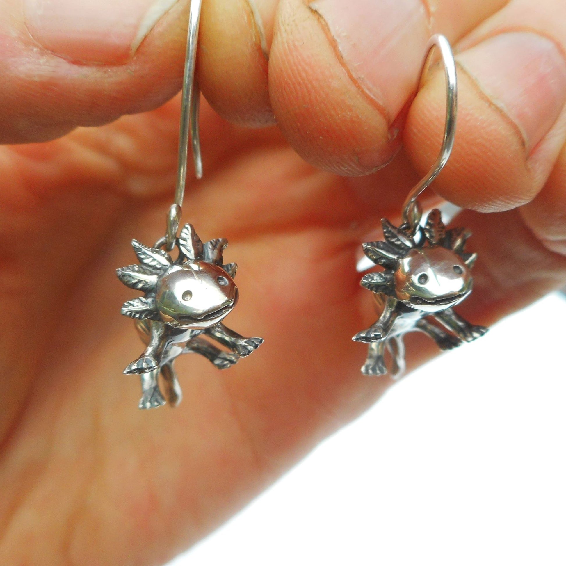 A photo of a pair of silver earrings in the shape of axolotls, held by a hand.