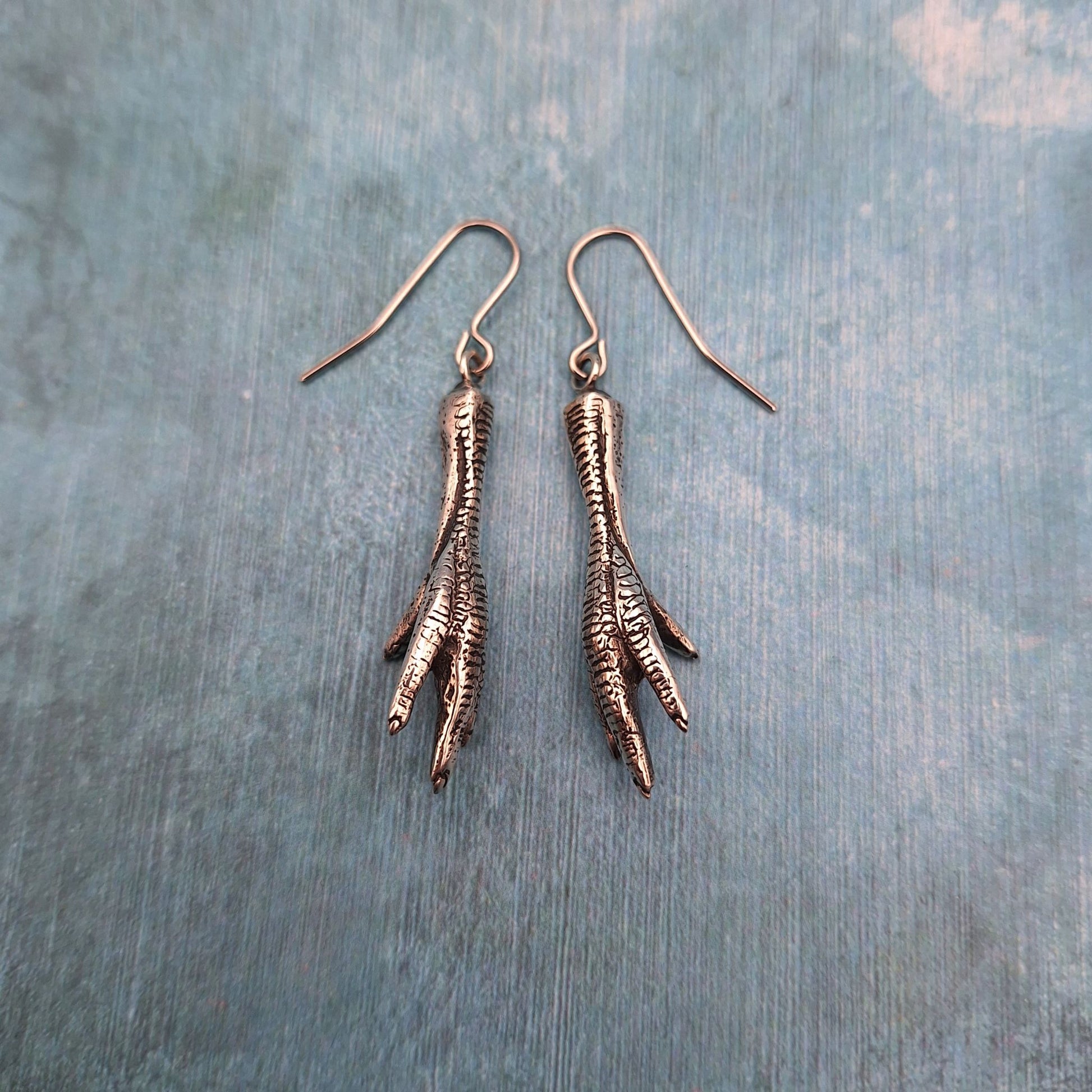 A photo of a pair of silver chicken foot earrings, on a blue background.