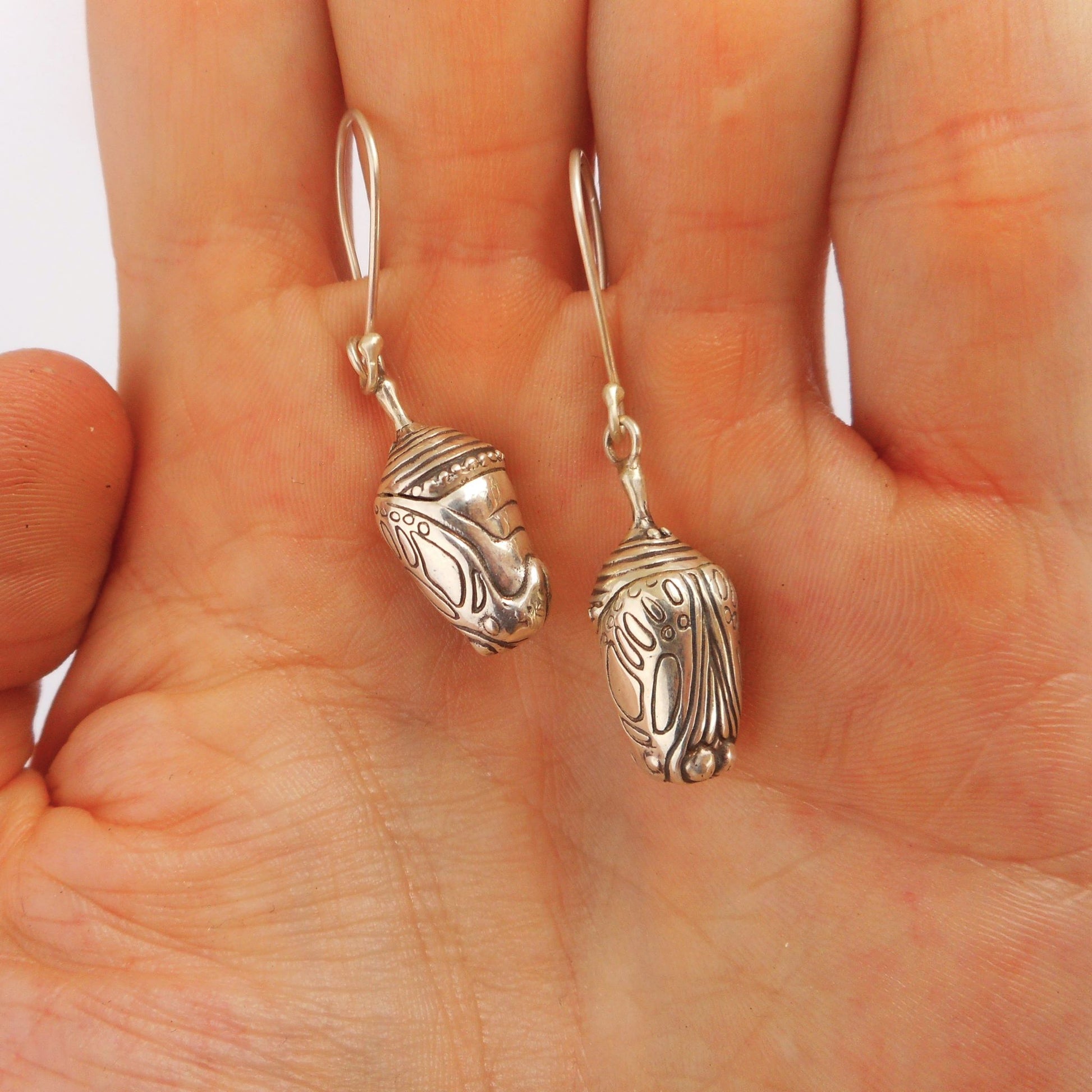 A photo of a pair of earrings n the shape of chrysalises, made of silver, hanging from the palm of a hand.