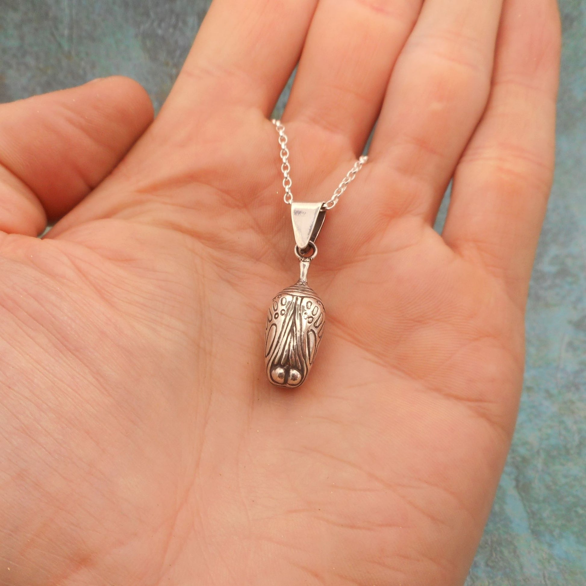A photo of a silver necklace shaped like a chrysalis, hanging on the palm of a hand, on a blue background.