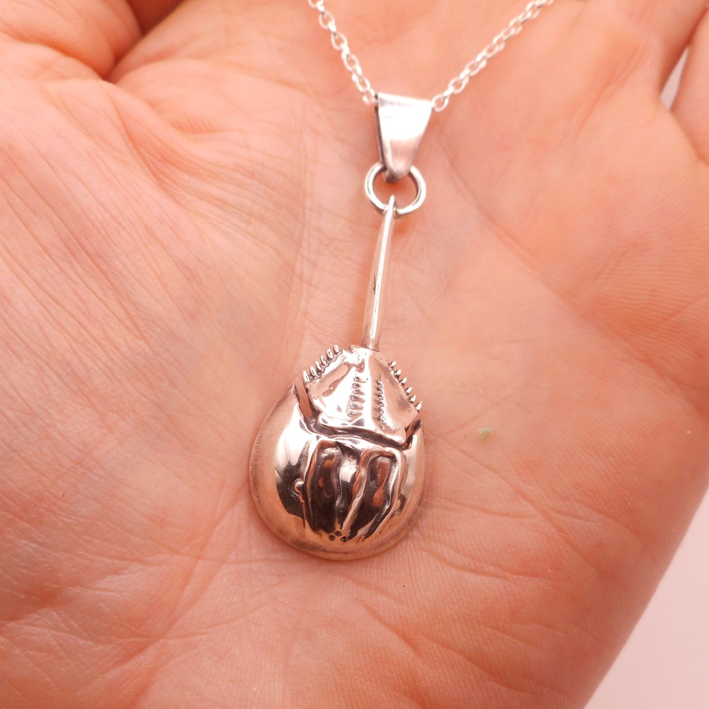 A photo of a silver pendant in the shape of a horseshoe crab, on the palm of a hand.
