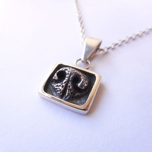 A photo of a silver pendant, shaped like a dog's nose in a square frame, on a white background.