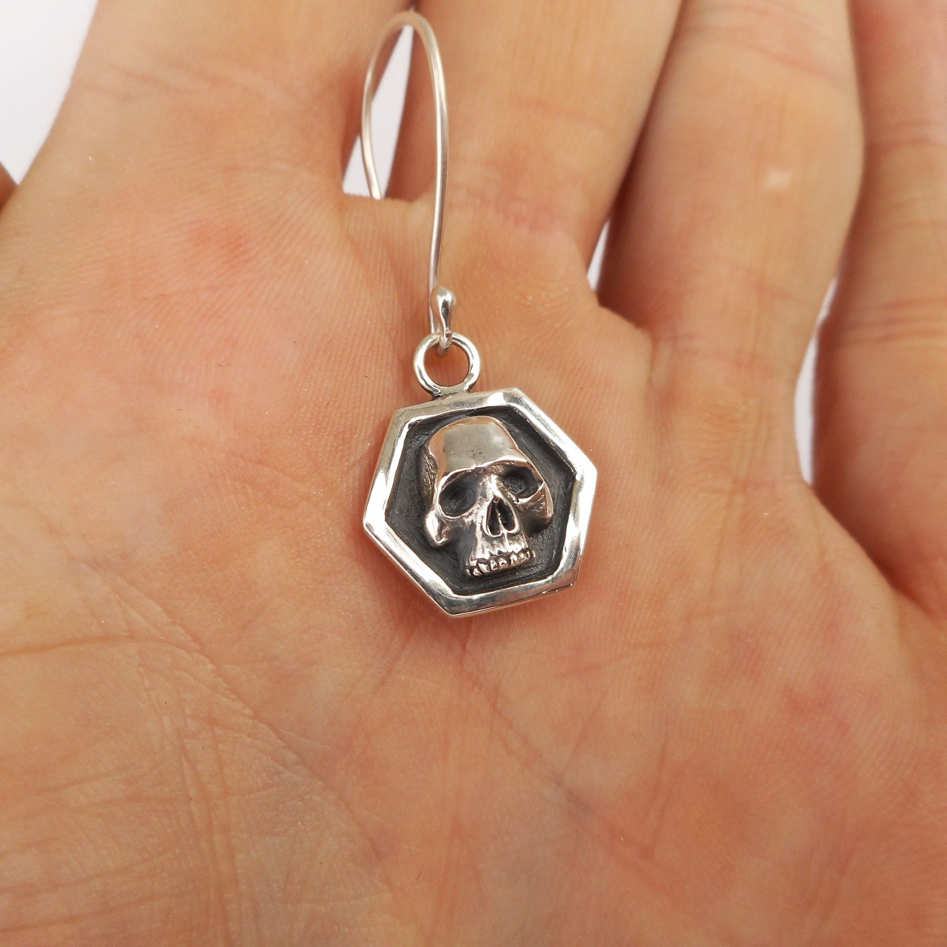 A photo of a silver earring in the form of a small skull in a hexagon, resting on the palm of a hand.