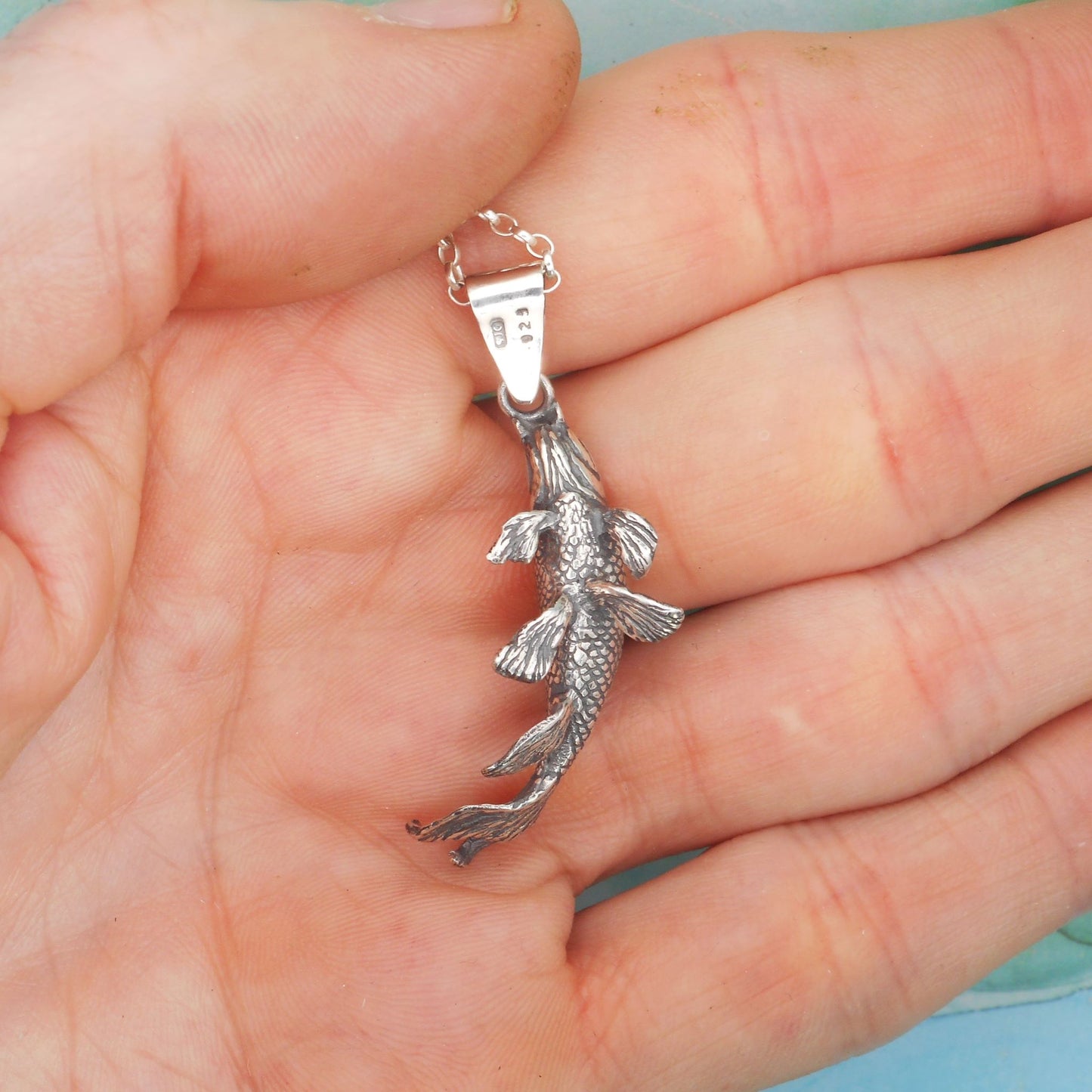 A photo of a silver necklace in the shape of a koi fish, resting on the palm of a hand.