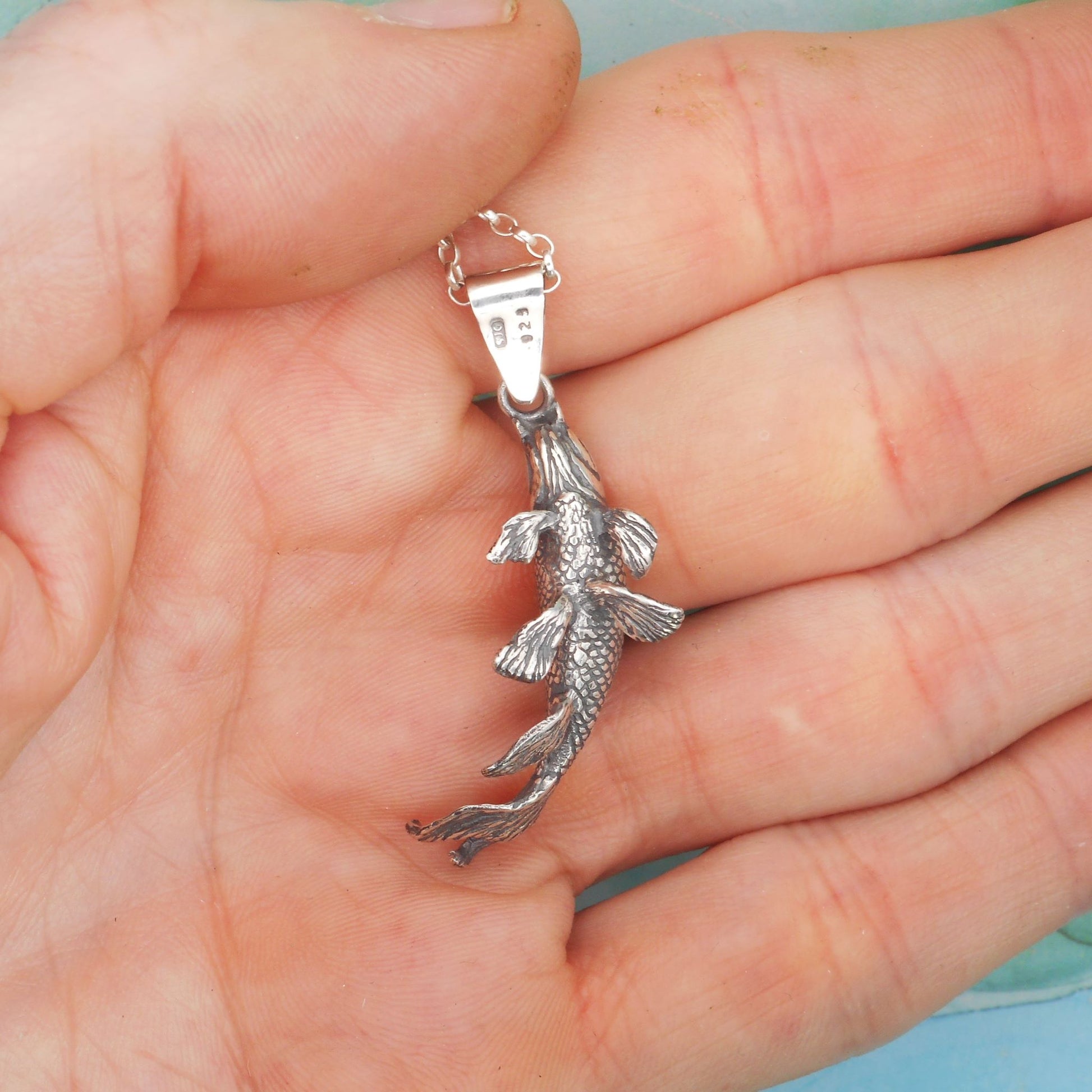 A photo of a silver necklace in the shape of a koi fish, resting on the palm of a hand.