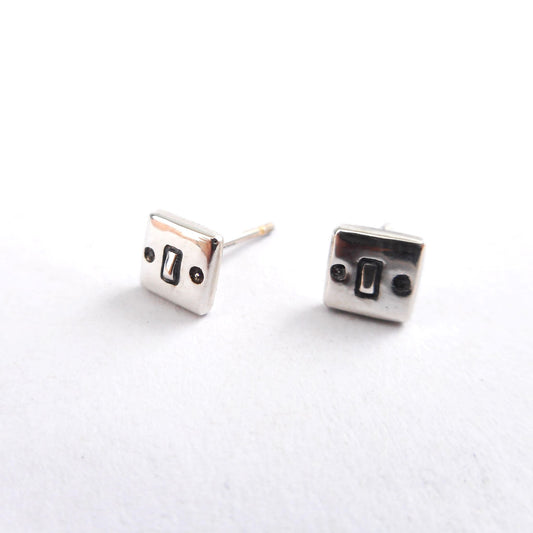 A photo of two silver ear studs shaped like miniature light switches.
