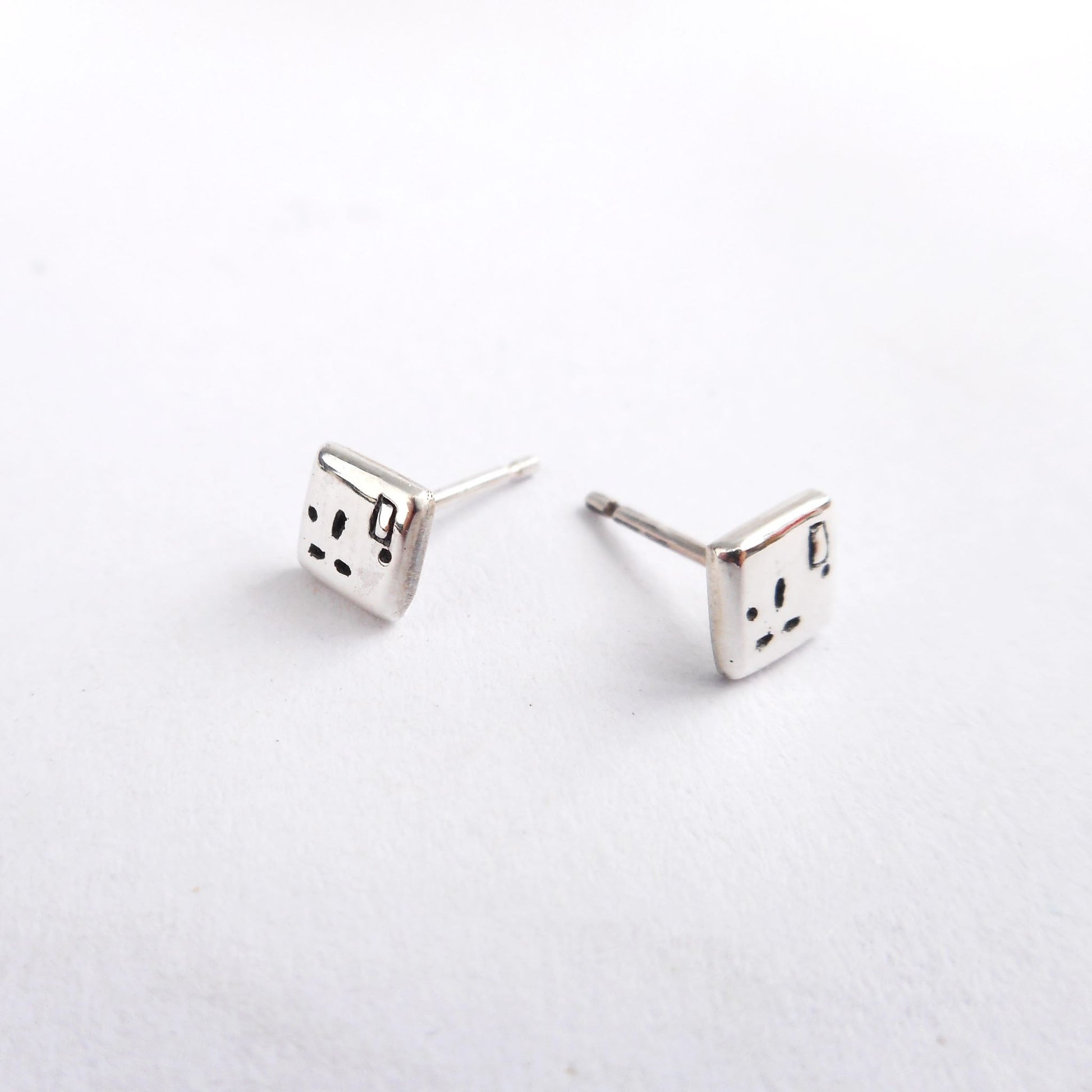 A photo of a pair of silver ear studs in the shape of plug sockets, on a white background.