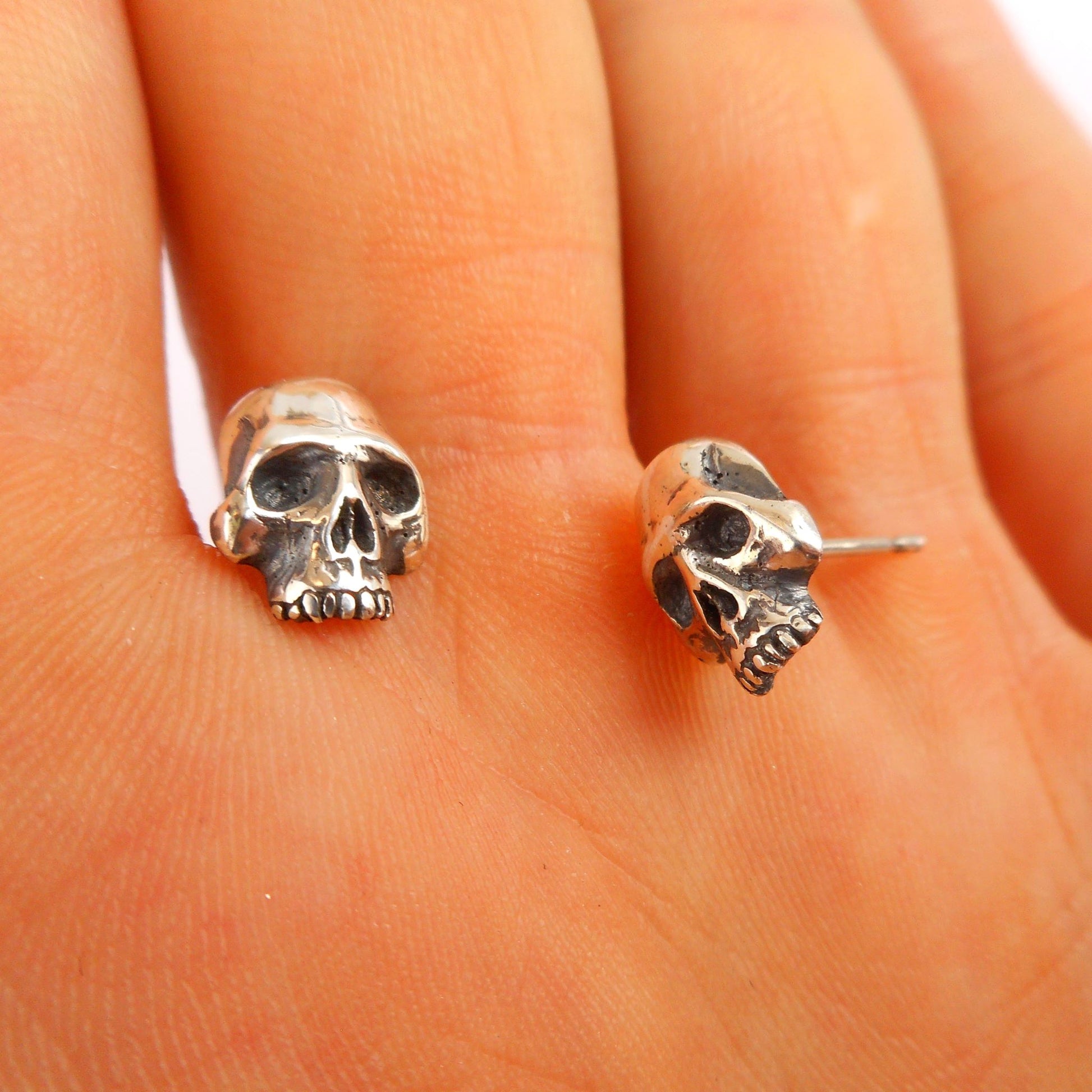 A photo of a pair of silver stud earrings shaped like small skulls, resting on the fingers of a hand.