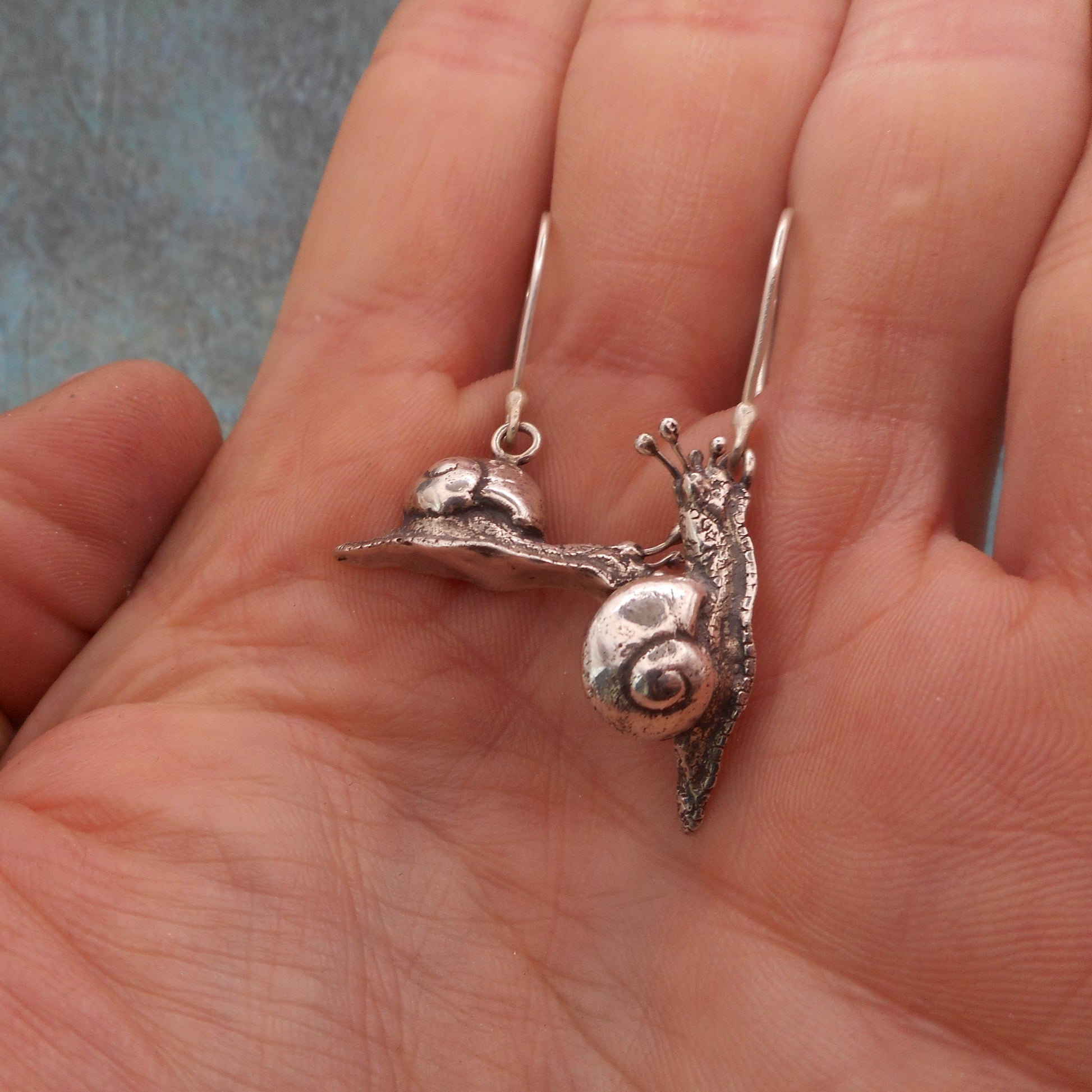 A photo of wo earrings in the shape of snails, resting on a hand.