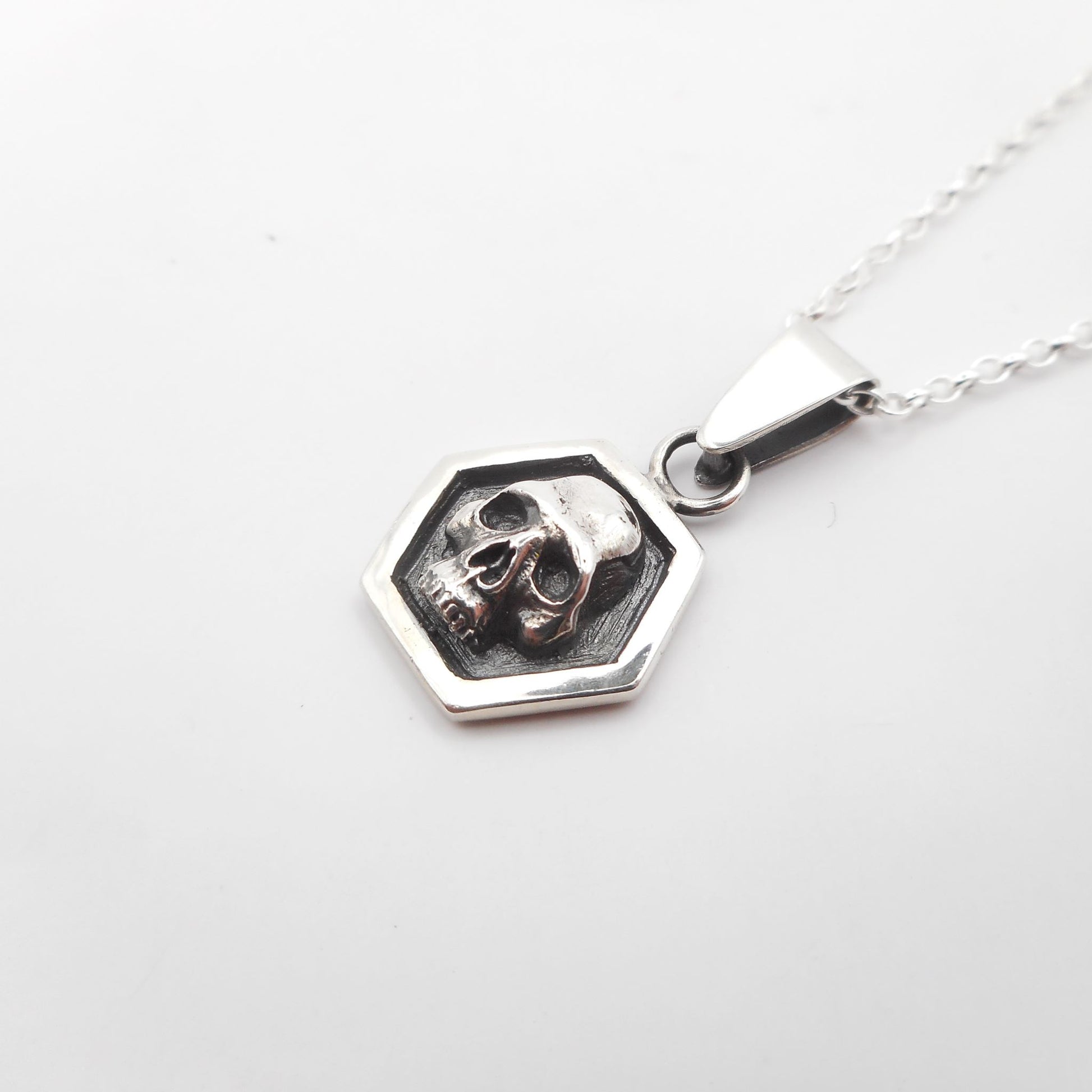 A photo of a small silver pendant in the shape of a skull surrounded by a hexagonal frame, resting on a white background.