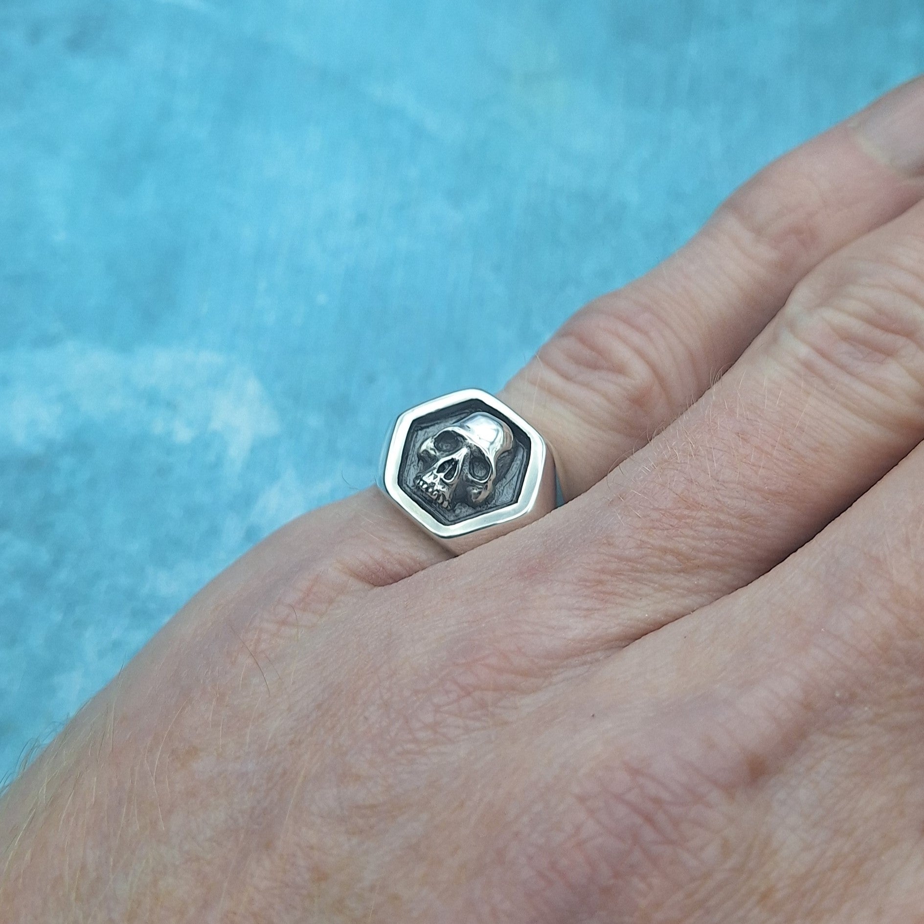 A photo of a silver skull ring, worn on the little finger of a hand, on a blue background.