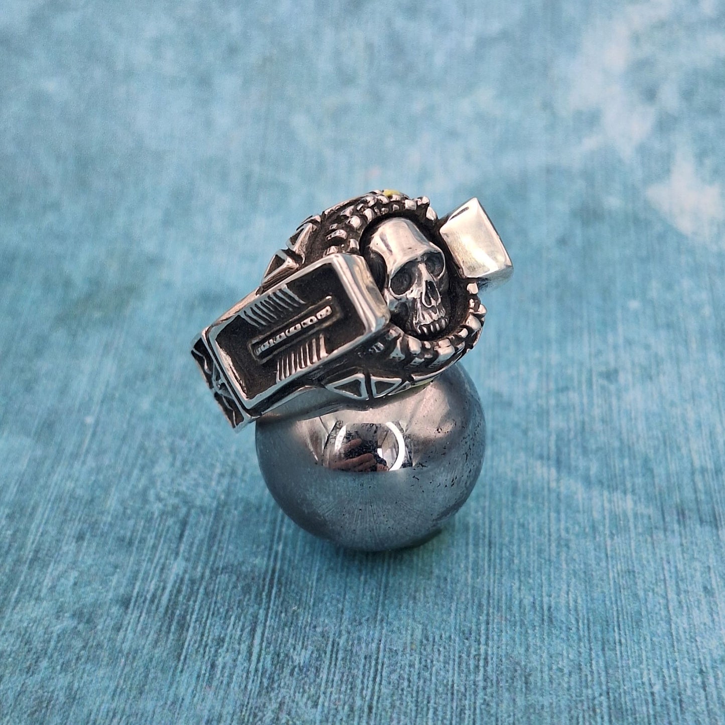 A photo of a silver skull ring, resting on a large ball bearing, on a blue background.