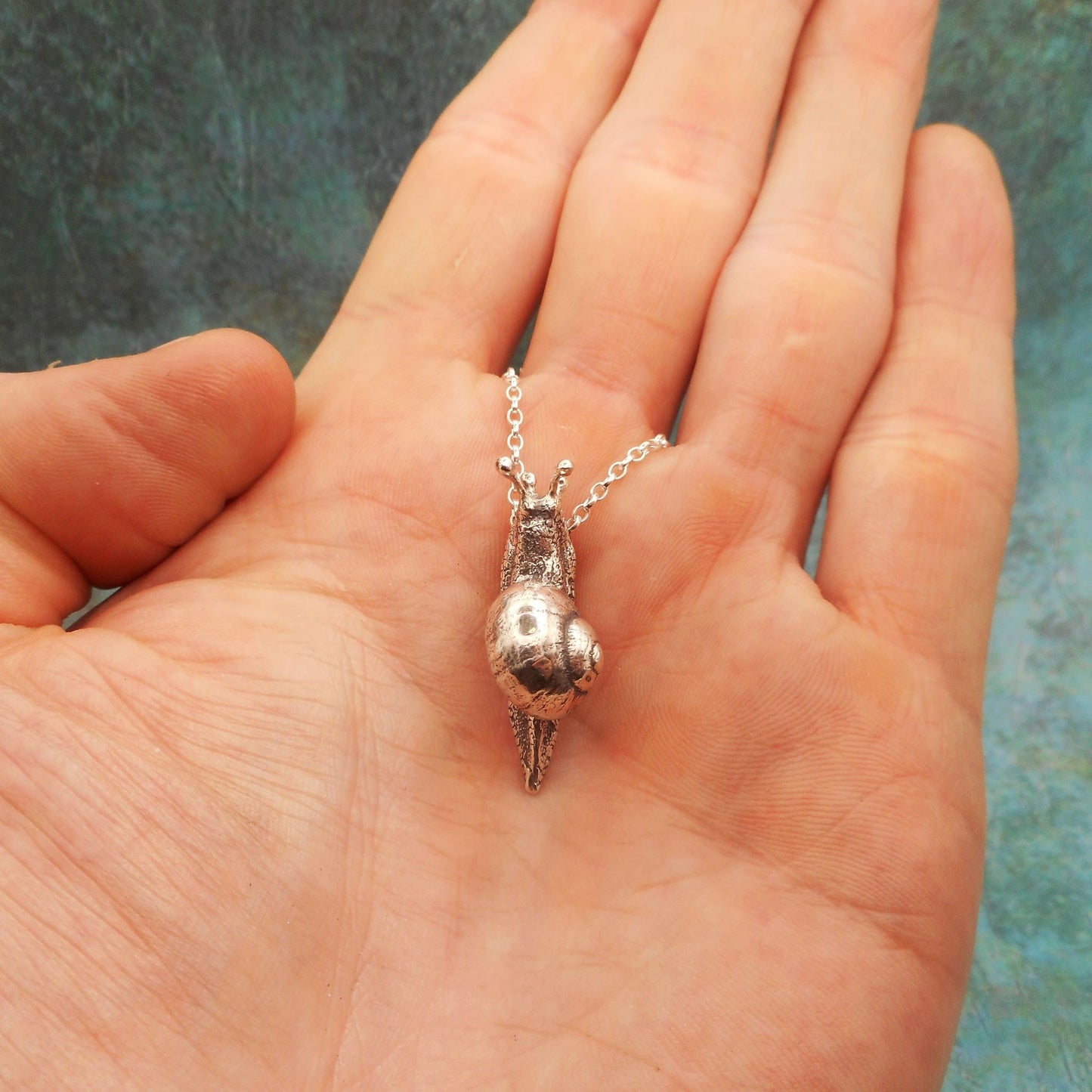 A photo of a silver snail necklace on the palm of a hand, against a blue background.