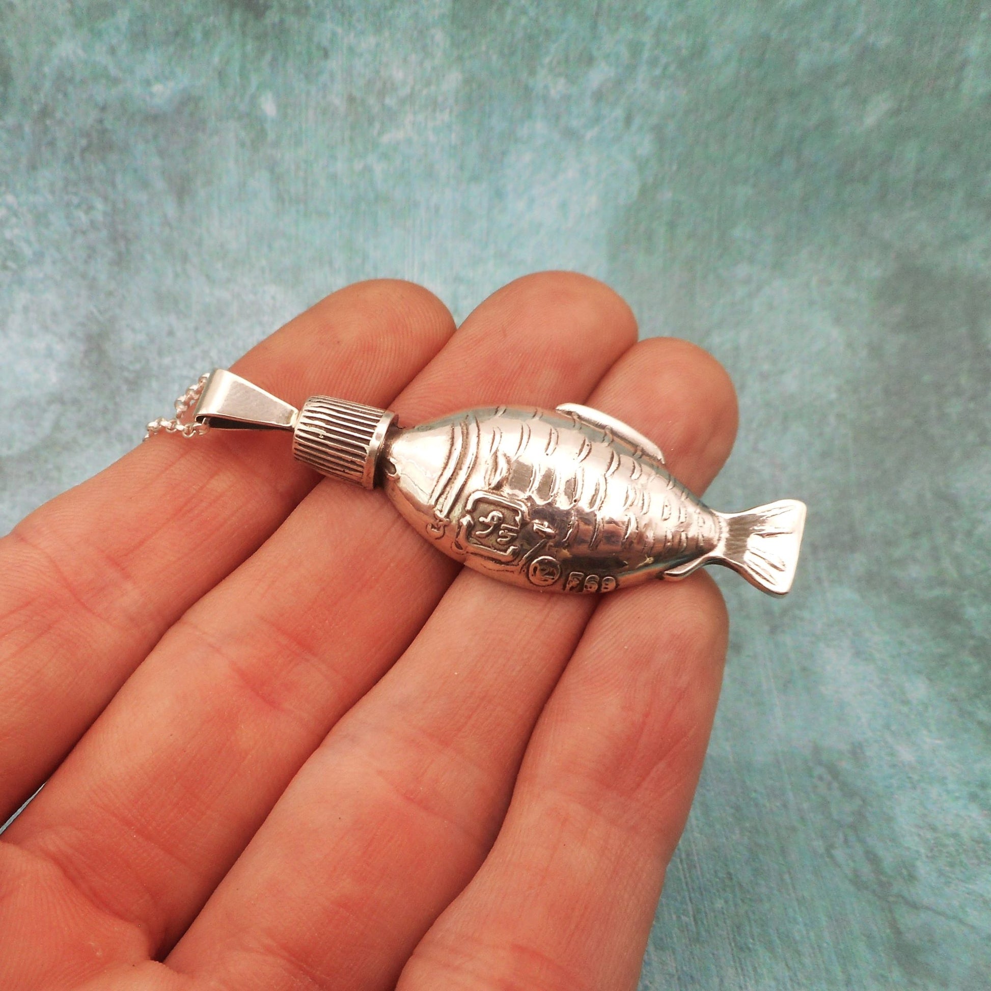 A photo of a silver necklace shaped like a soy sauce fish bottle, on the fingers of a hand, against a blue background.