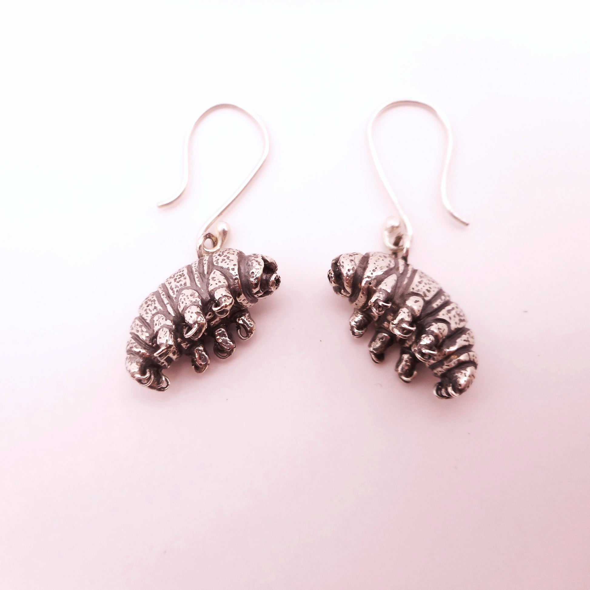 A photo of a pair of silver earrings shaped like tardigrades, on a white bakground.