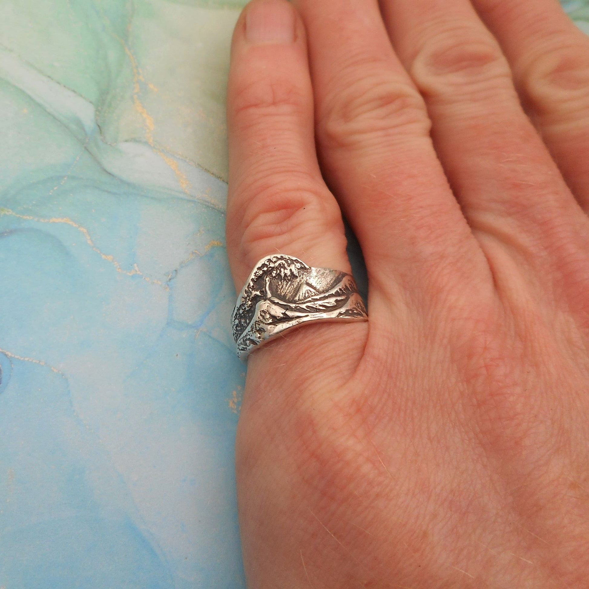 a photo of a silver ring which depicts an illustration of a wave, worn on a little finger.
